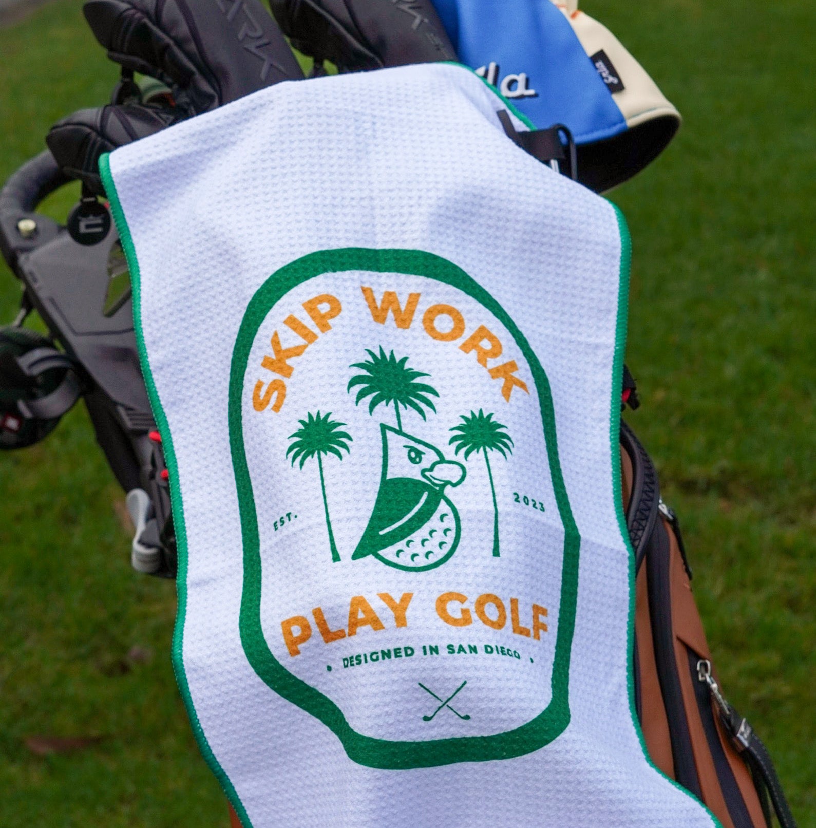 The Skip Work Golf Towel, featuring a bird, palm trees, and the text "SKIP WORK PLAY GOLF" in yellow and green, is draped over a golf bag on vibrant green grass.