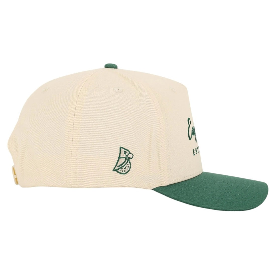The Heritage | Golf Hat is cream-colored with a green curved brim and features a small embroidered bird holding a golf ball on the left side. Made of premium cotton, it includes ventilation eyelets and an adjustable metal back strap.