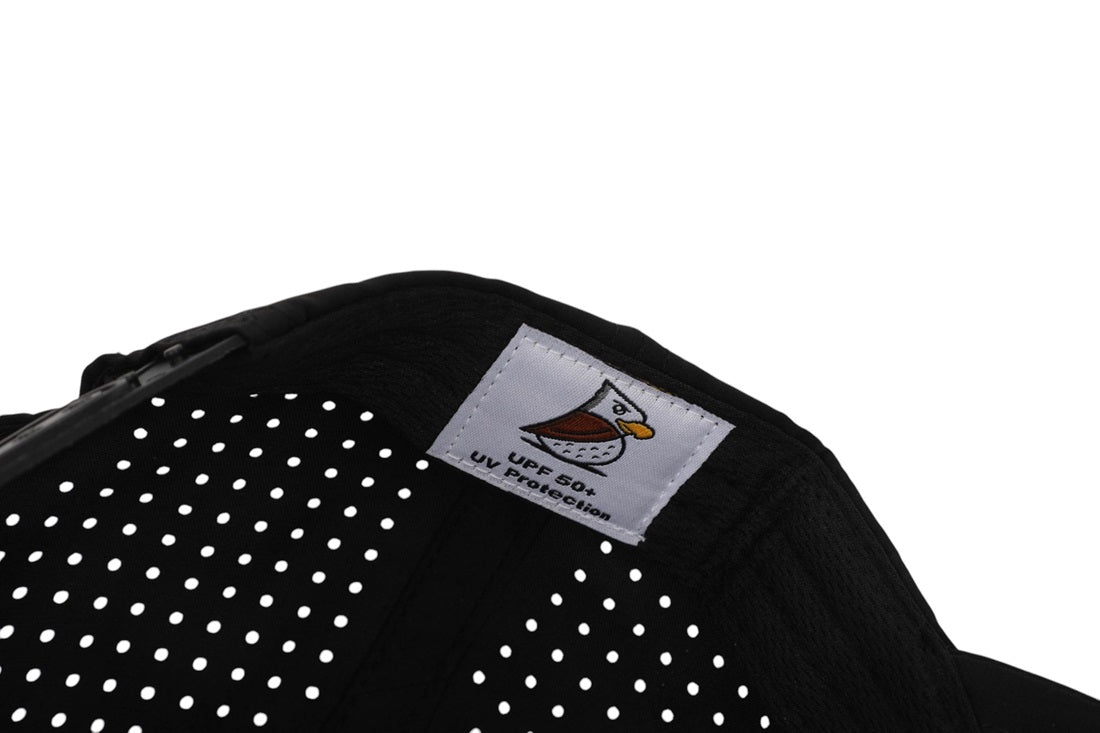 Close-up of the SL | Rope Golf Hat, black and made from moisture-wicking fabric with ventilation holes, a sewn-on white label with a cartoon bird, "UPF 50+ UV Protection" text, and a partially visible left-side zipper.