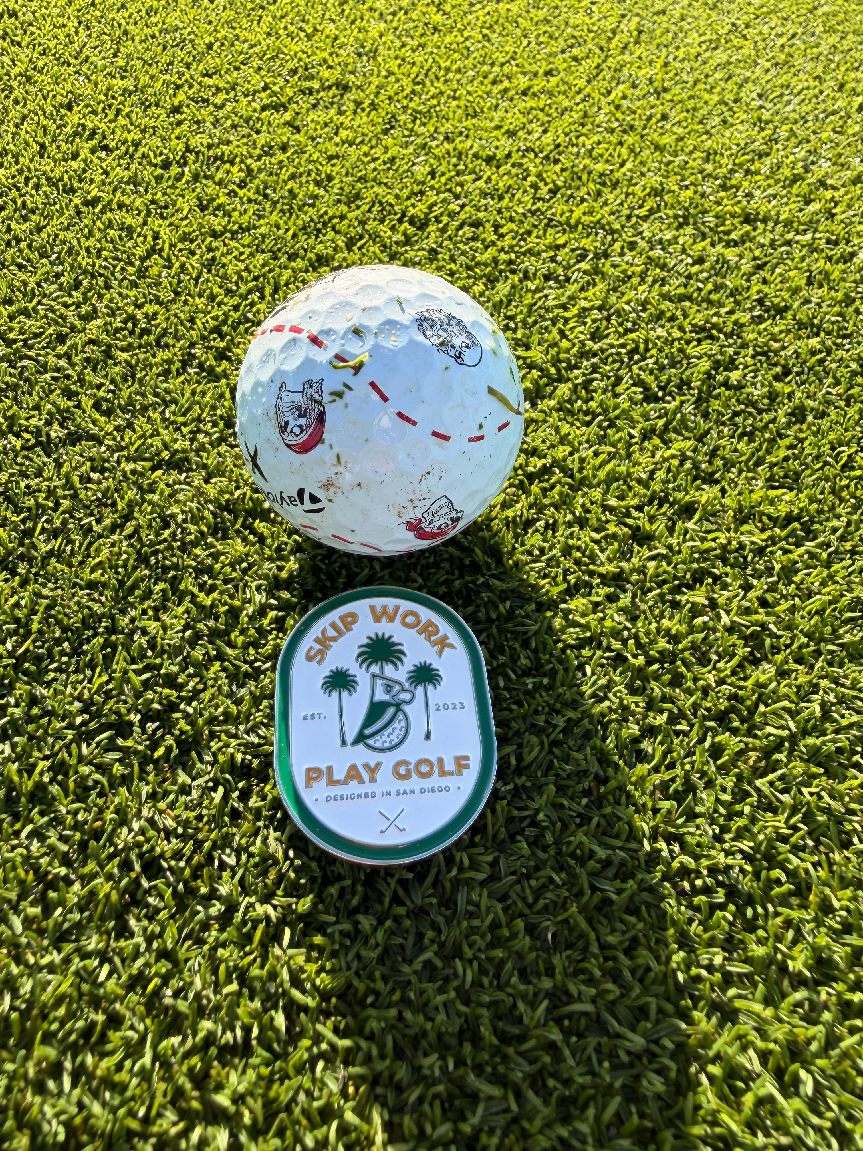 The Skip Work Bundle features a golf ball, sleek metal ball marker, and a round badge with "SKIP WORK PLAY GOLF" and palm trees, displayed on bright green artificial turf in the sunlight.
