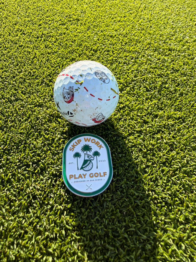The Skip Work Bundle features a golf ball, sleek metal ball marker, and a round badge with "SKIP WORK PLAY GOLF" and palm trees, displayed on bright green artificial turf in the sunlight.