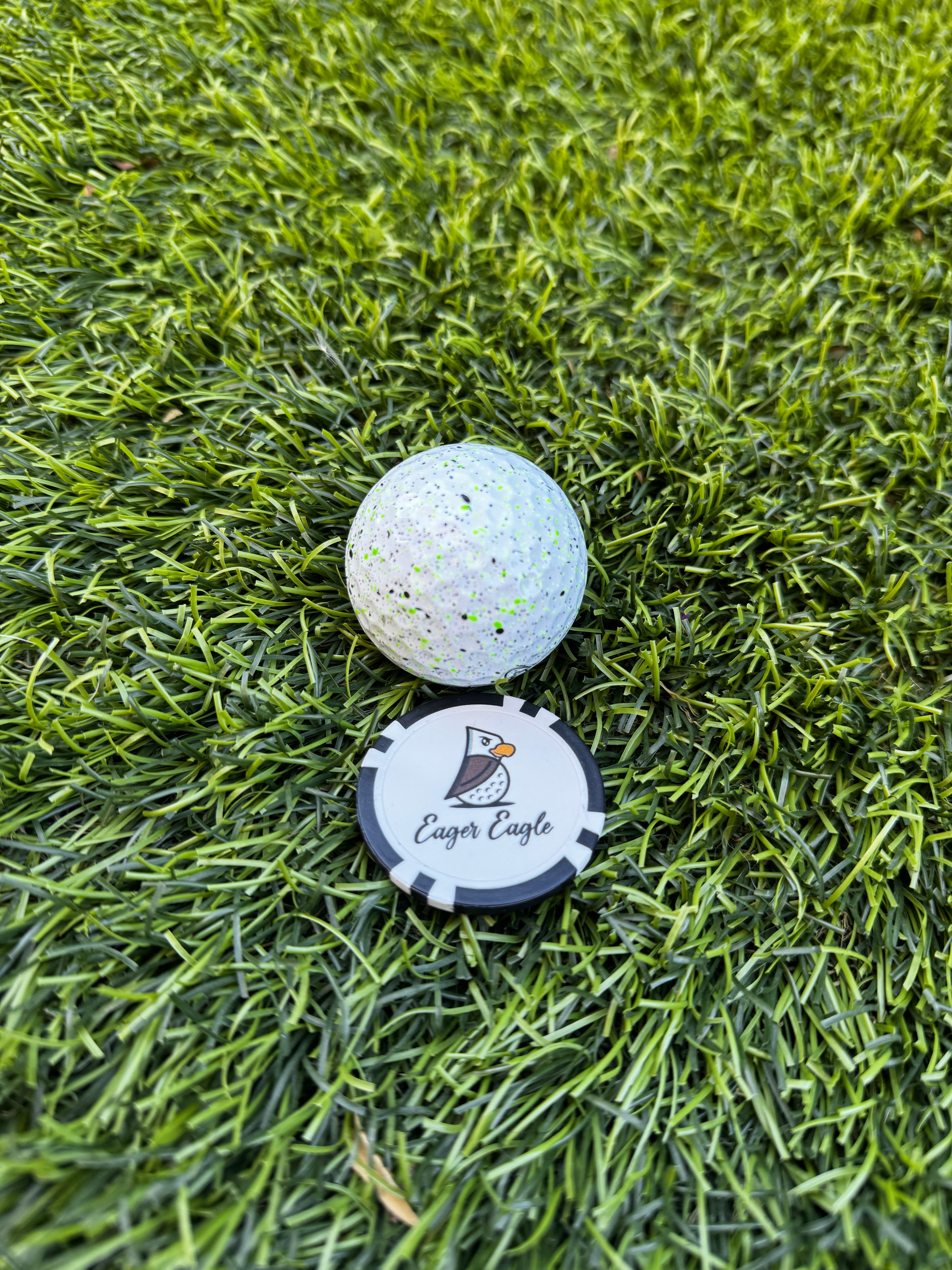 An Eager Eagle Poker Chip Ball Marker and a white golf ball with black speckles rest on green grass.