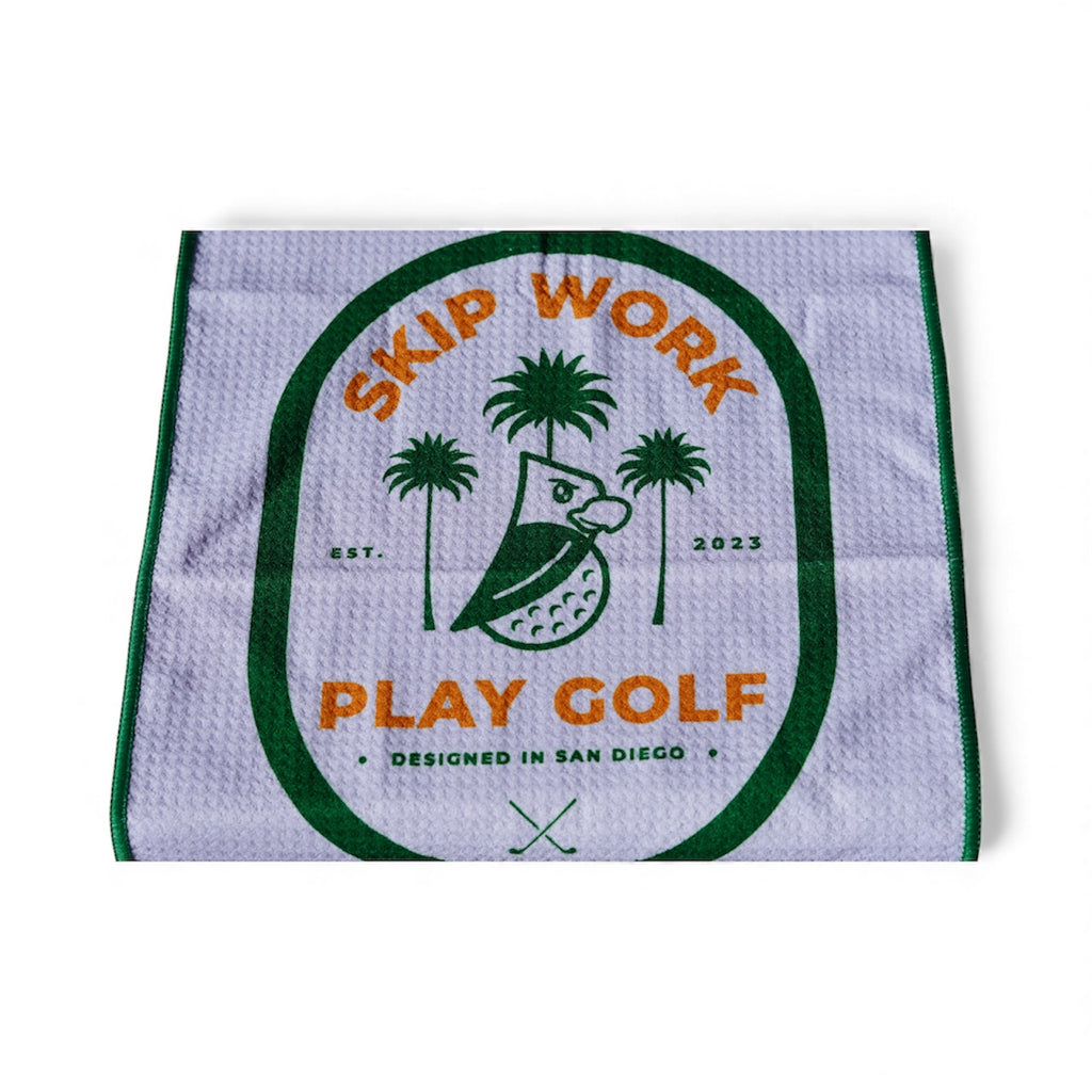 The Skip Work Bundle includes a white waffle golf towel with green and orange trim featuring an owl, palm trees, “SKIP WORK PLAY GOLF,” two crossed clubs, “EST. 2023,” and “DESIGNED IN SAN DIEGO.”.