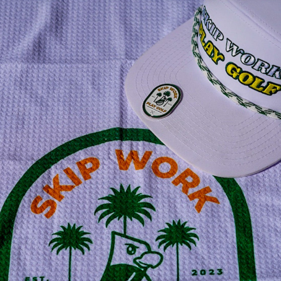 The Skip Work Bundle includes a lavender waffle golf towel with green border featuring a cartoon duck, palm trees, and "SKIP WORK," plus a white golf hat with "SKIP WORK PLAY GOLF" embroidered and a green braided rope above the brim.