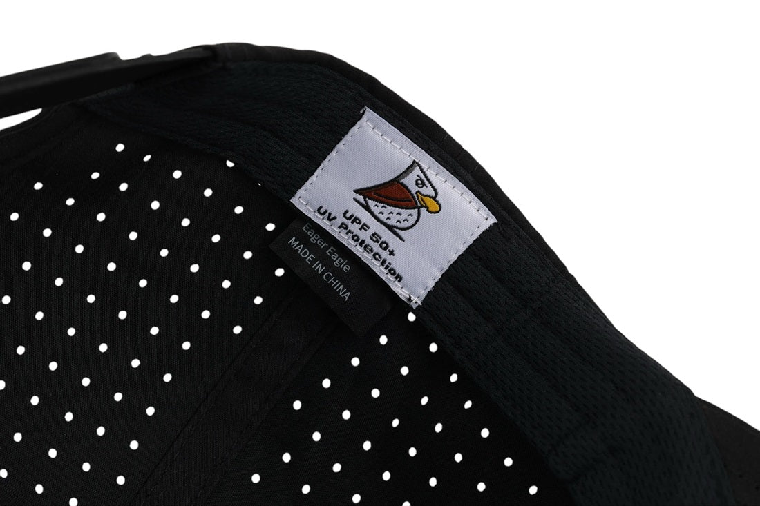 Close-up of Stealth | Golf Hat’s black fabric with white polka dots, a white Eager Eagle Golf Hats label with bird logo, UPF 50+ UV Protection, and MADE IN CHINA tag—all crafted from moisture-wicking material for all-day comfort.